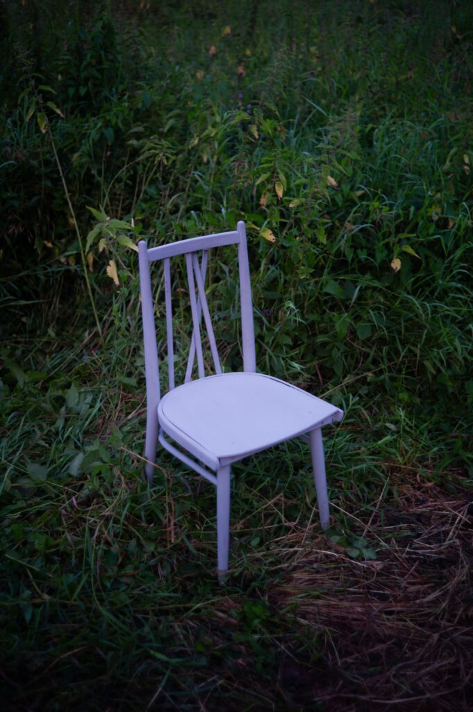 A solitary white wooden chair amidst lush overgrown greenery in a garden.